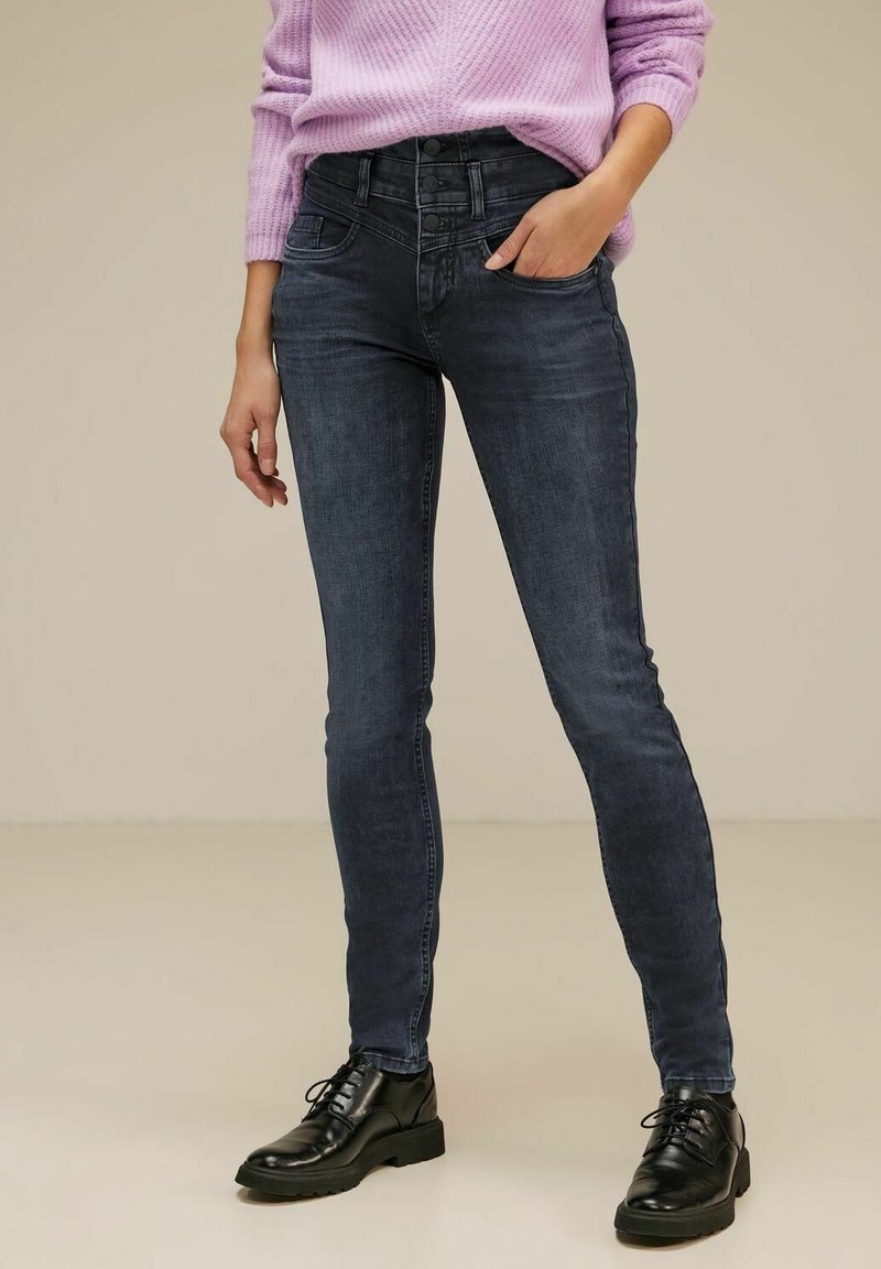 Femme portant un jean skinny taille haute foncé avec fermeture à triple bouton, un pull lavande et des chaussures à lacets noires, debout avec une main dans la poche.