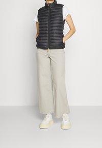 Chaleco negro acolchado con cuello alto, cierre de cremallera y tirador de cremallera naranja. Combinado con pantalones de pierna ancha de color beige claro y zapatillas blancas.