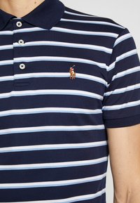 Námořnická modrá polo tričko se střídajícími světle modrými pruhy. Má klasický límec, tři knoflíky a malé vyšívané logo na hrudi.