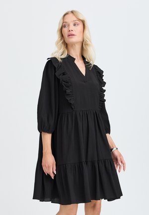 Femme blonde portant une robe noire longueur genou avec des détails à volants sur le devant et des manches trois-quarts, debout devant un fond uni.
