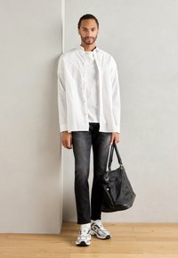 Cămașă albă cu dungi, purtată peste un tricou alb, blugi negri și adidași gri. Geantă neagră cu mânere. Podea din lemn și fundal deschis.