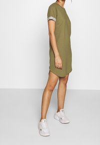 Oliwkowa sukienka z krótkim rękawem, okrągłym dekoltem i zgiętymi jasnoszarymi mankietami; miękki materiał, lekko zaokrąglony dół, noszona z białymi trampkami.