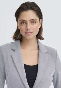 Blazer gris texturé avec des revers structurés, doté d'une fermeture à un seul bouton et porté sur un haut noir. Finition en tissu lisse.