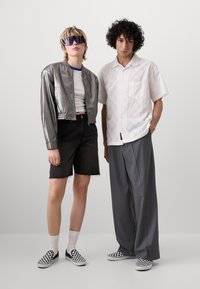 Strieborná metalická bomber bunda, biely crop top, čierne džínsy a šachovnicové slip-on topánky; muž v bielej vzorovanej košeli a šedých nohaviciach.