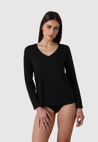 Donna con lunghi capelli scuri che indossa un top nero a maniche lunghe con scollatura a V e biancheria intima nera, in piedi contro uno sfondo chiaro e semplice.