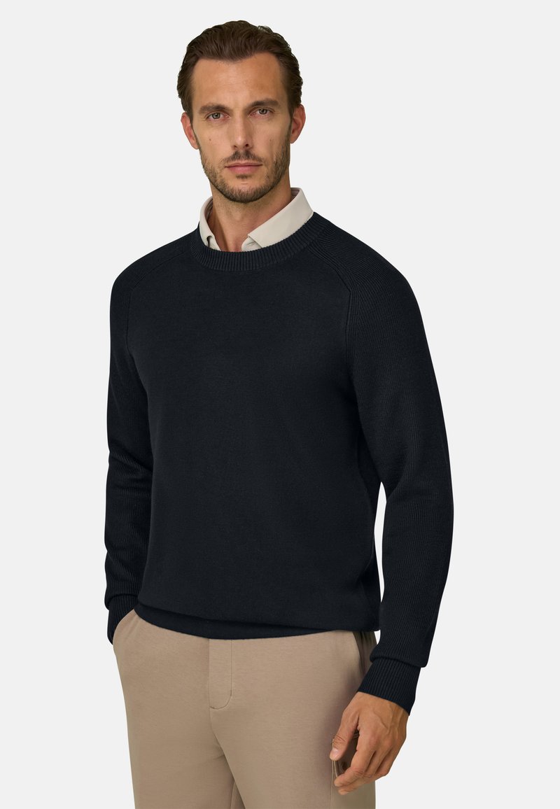 Homme portant un pull noir à col rond sur une chemise blanche à col et un pantalon beige, debout avec une main dans la poche devant un fond uni.