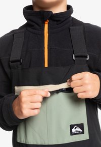 Svart fleece-topp med orange dragkedja, justerbara svarta remmar och grön ficka. Inkluderar logotypmärke längst ner till höger. Texturen är mjuk.