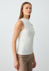 Top senza maniche lavorato a maglia in bianco con colletto alto, caratterizzato da perle decorative distribuite uniformemente e dettagli metallici. Vestibilità slim.
