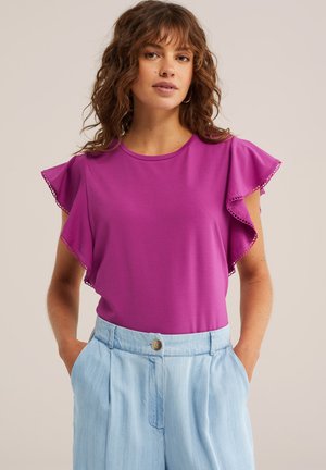 Vrouw met krullend haar die een paarse top met vleermuismouwen en lichtblauwe, hooggetailleerde broek draagt, met haar handen in de zakken.