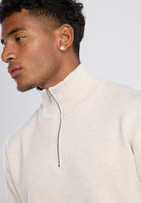 Beige zip-up sweater with a high collar, soft texture, and a metallic zipper. Features minimal stitching and a fitted design.