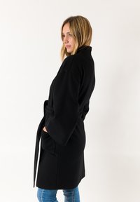 Roba nera con vita cinturata, maniche ampie e tasche laterali. Tessuto morbido con un design minimale, che offre una vestibilità rilassata e una texture liscia.