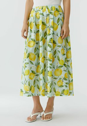 Jupe midi fluide à imprimé citron jaune et feuilles vertes, portée avec des sandales blanches à talons et un top blanc.