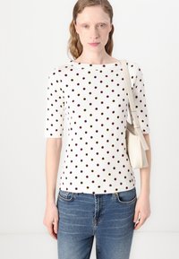 Haut blanc à manches courtes avec des pois rouges et noirs. Il présente un col bateau et est associé à un jean en denim bleu et un sac à main clair.