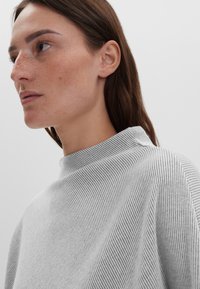 Graues, geripptes Strickoberteil mit hohem Kragen, das über eine weiche Textur und eine lockere Passform verfügt. Der Stoff zeigt subtile Linien und kontrastiert mit dem Hintergrund.