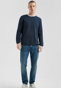 Marineblauw gebreid shirt met lange mouwen en een ronde hals, gecombineerd met lichtblauwe jeans. Het model draagt witte sneakers voor een casual look.
