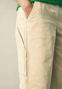 Pantalons cargo beiges avec poches latérales, fabriqués à partir d'un tissu doux ; dotés d'un devant plat, de coutures renforcées et d'une coupe décontractée.