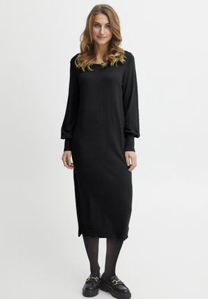Robe en tricot noire à manches longues avec un col rond et des fentes latérales ; portée avec des chaussures noires épaisses présentant des accents en métal doré.