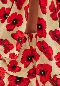 Robe à fleurs avec un fond crème, ornée de grands motifs de coquelicots rouges et de centres noirs, en tissu texturé, et avec un détail de fermeture éclair discret.