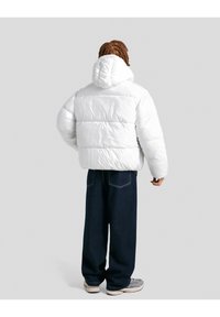 Jachetă albă tip puf, cu glugă, având secțiuni captusite și o textură strălucitoare, purtată peste blugi largi de un albastru închis și adidași gri.