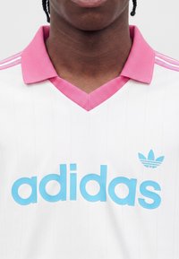 Άτομο που φοράει λευκό μπλουζάκι Adidas με ροζ γιακά και ανοιχτό μπλε λογότυπο, εστιάζοντας στον λαιμό και την ανώτερη περιοχή του στήθους.