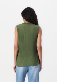 Femme aux longs cheveux bouclés portant un haut sans manches côtelé vert olive et un jean bleu clair, debout devant un fond blanc uni.