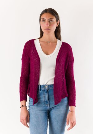 Donna con capelli lunghi e scuri indossa un cardigan rosa scuro strutturato sopra una maglietta bianca a scollo a V e jeans blu, in piedi davanti a uno sfondo semplice.