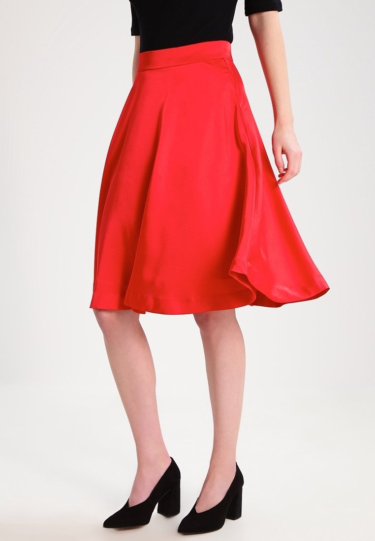 Femme portant une jupe midi rouge, un haut noir et des chaussures noires à talons carrés, debout devant un fond blanc uni.