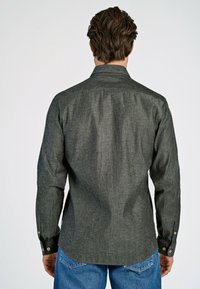 Bakvy av en mörkgrå, texturerad skjorta med knapp-up med långa ärmar och knappdetaljer vid manschetterna, i kombination med ljusblå jeans.