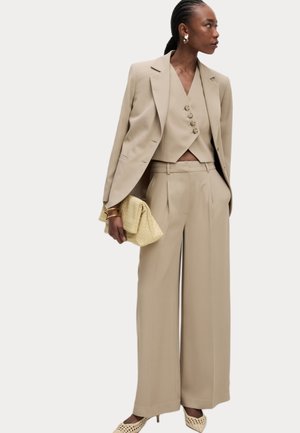 Ensemble de costume beige sur mesure avec un gilet ajusté à double boutonnage, un pantalon large taille haute et des escarpins assortis à bout pointu. Sac à main jaune.