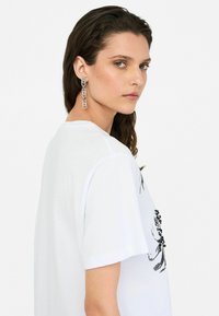 Tricou alb din bumbac cu un design floral negru imprimat. Modelul poartă cercei din argint în formă de lanț, arătând un profil lateral și o textură netedă.