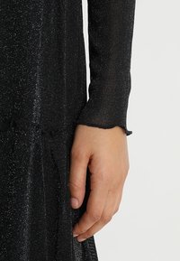 Black dress with a fitted, long-sleeve design. Made from textured fabric with metallic threads, featuring a frayed cuff and flared hem.