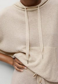Sudadera con capucha beige de textura suave, mangas cortas, cordones y un bolsillo frontal. Diseño simple con acentos metálicos mínimos.