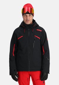 Giacca da sci nera con dettagli rossi, tasche con zip e cappuccio regolabile. Indossata con pantaloni rossi e guanti neri, completata da un berretto rosso acceso e occhialoni.