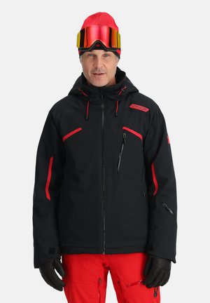 Black ski jacket with red accents, zippered pockets, and adjustable hood. Worn with red pants and black gloves, complemented by a bright red beanie and goggles.