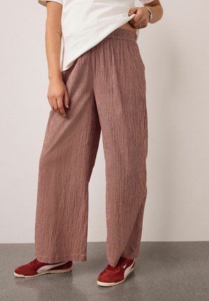 Frau trägt locker sitzende, rötlich karierte Hose mit weitem Bein, leicht hochgezogenes weißes Hemd und rote Turnschuhe, steht auf grauem Boden.