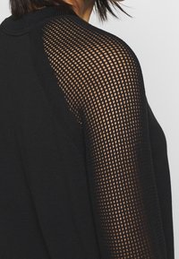 Czarny sweter o dopasowanej sylwetce z teksturowanym siatkowym ramieniem. Siatka ma wzór w postaci siatki, co dodaje wizualnego kontrastu do materiału.