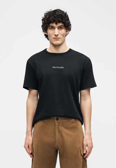 Νεαρός άνδρας με σγουρά μαλλιά που φοράει μαύρο μπλουζάκι με το κείμενο "The Kooples" και καφέ παντελόνι, στέκεται μπροστά σε απλό φόντο.