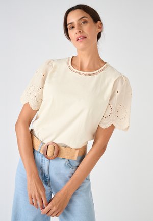 Donna che indossa una blusa color crema con maniche corte di pizzo e uno scollo decorativo, abbinata a jeans azzurri chiari e una cintura beige intrecciata.