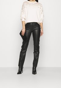 Lichtbeige sweatshirt met kantdetail op de schouders, gecombineerd met zwarte leren broek en zwarte enkellaarsjes met hak, houdend een zwarte clutch vast.