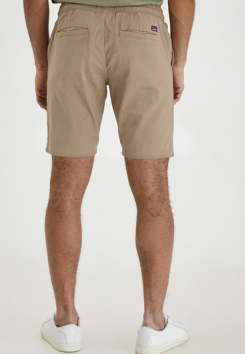 Homme portant un short beige avec des poches arrière et des baskets blanches, debout devant un fond clair uni.
