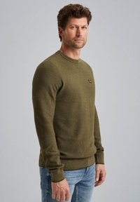 Olivovo zelený pletený sveter s textúrovanými pruhmi, okrúhlym výstrihom a akcentom loga. Dlhé rukávy a rebrovaný lem dodávajú detail. Nosený s modrými džínami.