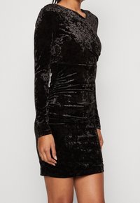 Robe longue à manches longues en velours noir avec une silhouette ajustée. Le tissu présente une texture douce et brillante ainsi qu'un léger froncé sur les côtés.