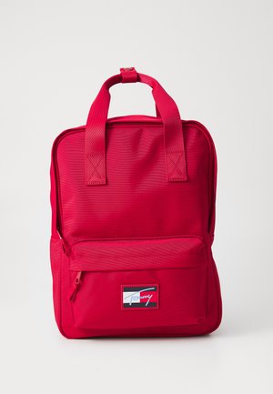Sac à dos en nylon rouge avec deux poignées, une poche zippée à l'avant et un écusson avec logo. Présente une forme structurée et une texture lisse.