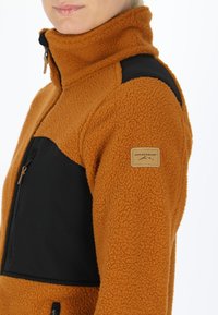 Giacca in pile arancione con accenti in tessuto nero, colletto alto e tasca con zip frontale. Presenta una toppa con logo sulla manica sinistra.