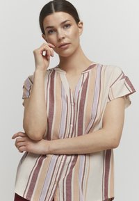 Chemise à manches courtes avec un boutonnage, présentant des rayures verticales dans des teintes de mauve, pêche et crème. Tissu doux et design en V.