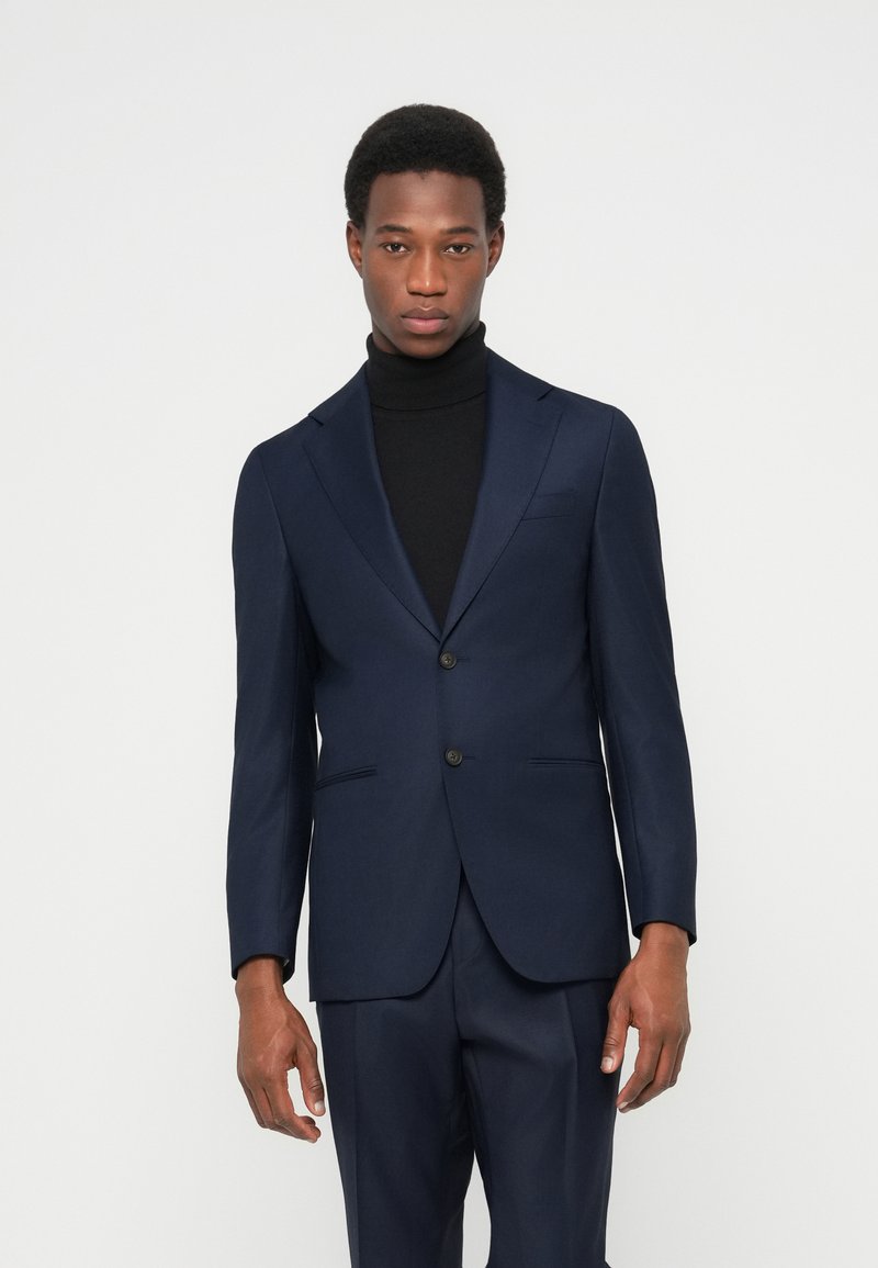 Marineblaue Anzugjacke aus glattem Stoff mit einem Revers mit Kerbe, zwei Knöpfen und Seitentaschen. Wird über einem schwarzen Rollkragenpullover getragen.