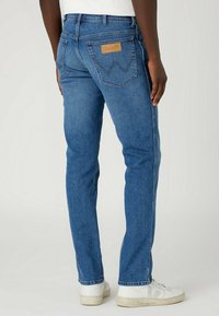 Pantalones vaqueros de mezclilla azul con un corte de pierna recta, que cuentan con un parche de cuero en el bolsillo trasero y zapatillas blancas. Detalles de costura visibles.