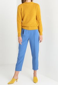 Senfgelber strukturierter Strickpullover, kombiniert mit hellblauen maßgeschneiderten Hosen. Das Model trägt gelbe Absatzschuhe. Einfacher Hintergrund.