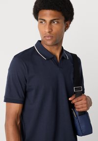 Ναυτικό πόλο μπλουζάκι με αντίθετο ανοιχτό περίγραμμα στο γιακά, διαθέτει κοντά μανίκια. Φοράει μια μαύρη τσάντα με λουρί κρατημένη στο δεξί χέρι.