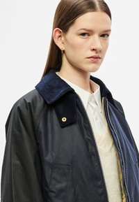 Tengerészkék dzseki kordbársony gallérral, cipzáras záródással és arany színű fémrészletekkel, világos színű pulóver és galléros ing fölött viselve. Simább textúra.